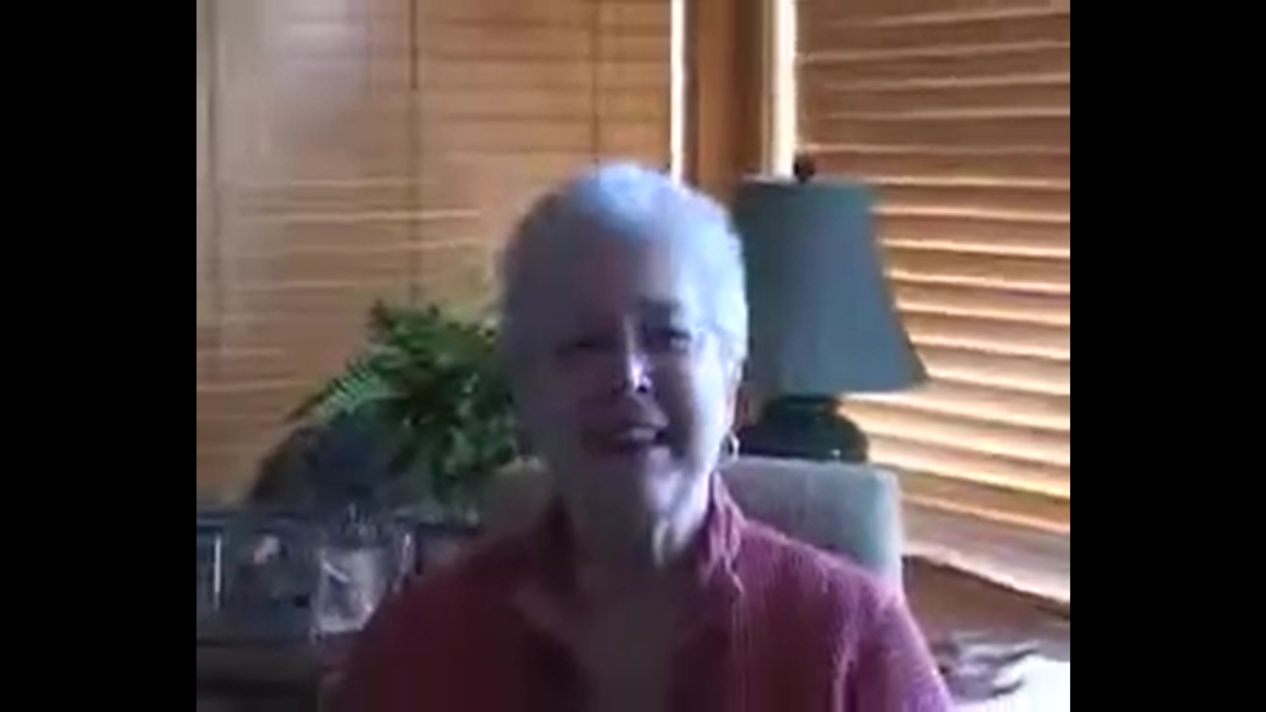Carol Kirkpatrick, an older woman with short white spiky hair, smiles at the camera in a dimly-lit room.